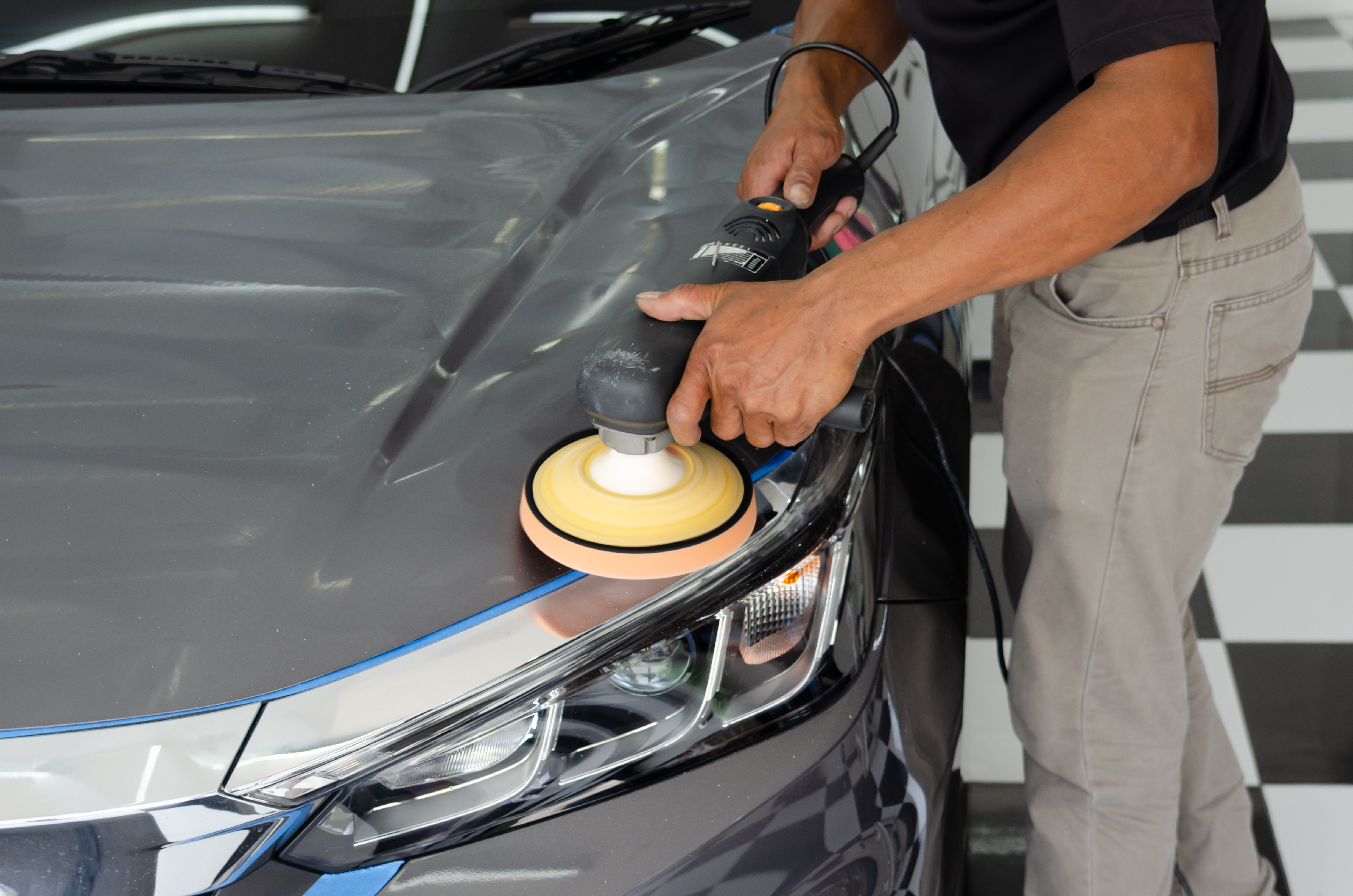 Professional detailer using orbital polisher with yellow foam pad on gray car hood